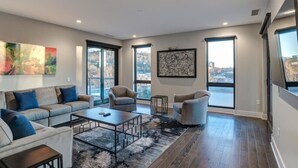 A spacious living room with a sofa set, a coffee table, and two armchairs. There are large windows providing a view of the city.