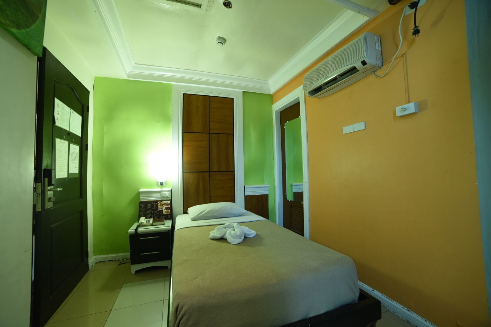 Habitación de hotel con una cama, una mesita de noche con teléfono, aire acondicionado de pared y una puerta con pared verde.