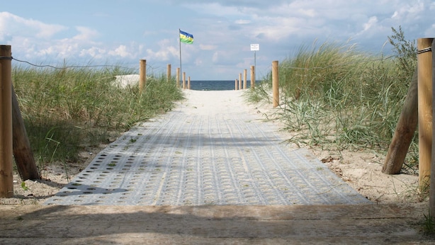 Un sentiero sabbioso conduce a una spiaggia, con una bandiera, pali di legno e un cielo sereno.
