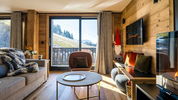 Un salon chaleureux avec un canapé, une table basse ronde et une cheminée. Une grande fenêtre offre une vue imprenable sur un paysage enneigé.