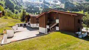 A wooden cabin with a staircase leading to a balcony, surrounded by a paved area and a view of snow-capped mountains. - Bright apartment, near slopes, pets allowed (La Clusaz)