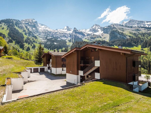 A wooden cabin with a staircase leading to a balcony, surrounded by a paved area and a view of snow-capped mountains.