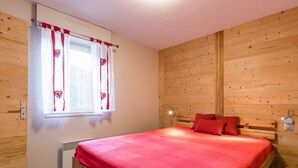 A wooden-paneled room with a bed, a window with curtains, a bedside lamp, and a thermostat. - Bright apartment, near slopes, pets allowed (La Clusaz)