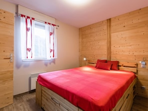 A wooden-paneled room with a bed, a window with curtains, a bedside lamp, and a thermostat.