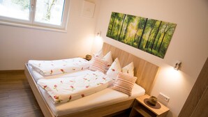 Un lit double avec une literie à motifs floraux, une table de chevet avec une lampe, un tableau représentant une forêt et une fenêtre donnant sur les arbres.