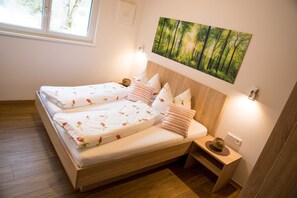 A double bed with floral bedding, a nightstand with a lamp, a painting of a forest, and a window with a view of trees. - Gaichtapart - Alpenapart Singer (Ehenbichl)