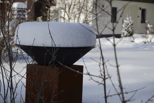 A snow-covered object on a pedestal with bare branches in the foreground and a building in the background. - Gaichtapart - Alpenapart Singer (Ehenbichl)