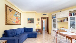 Sala de estar con un sofá azul, una mesa de comedor con sillas, una zona de cocina con armarios y un cuadro en la pared.
