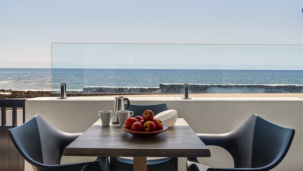 Et bord med frugt i en skål, en kaffekande og kopper, med udsigt over havet.