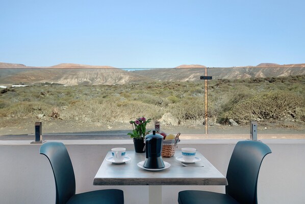 A table with a coffee maker, cups, and a basket of fruit, set against a desert landscape view.