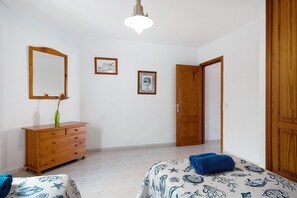 A bedroom with a wooden dresser, a bed with a blue and white patterned bedspread, a wooden door, and a wall-mounted mirror.