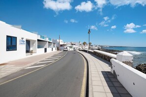 A coastal road with a white building on the left, a clear sky, and the ocean in the background.