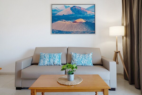 A living room with a sofa, two patterned pillows, a wooden coffee table with a plant, a lamp, and a framed landscape painting on the wall.