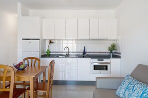 A compact kitchen with white cabinets, a dining table with chairs, a built-in oven, and a small seating area with a sofa and a patterned pillow.
