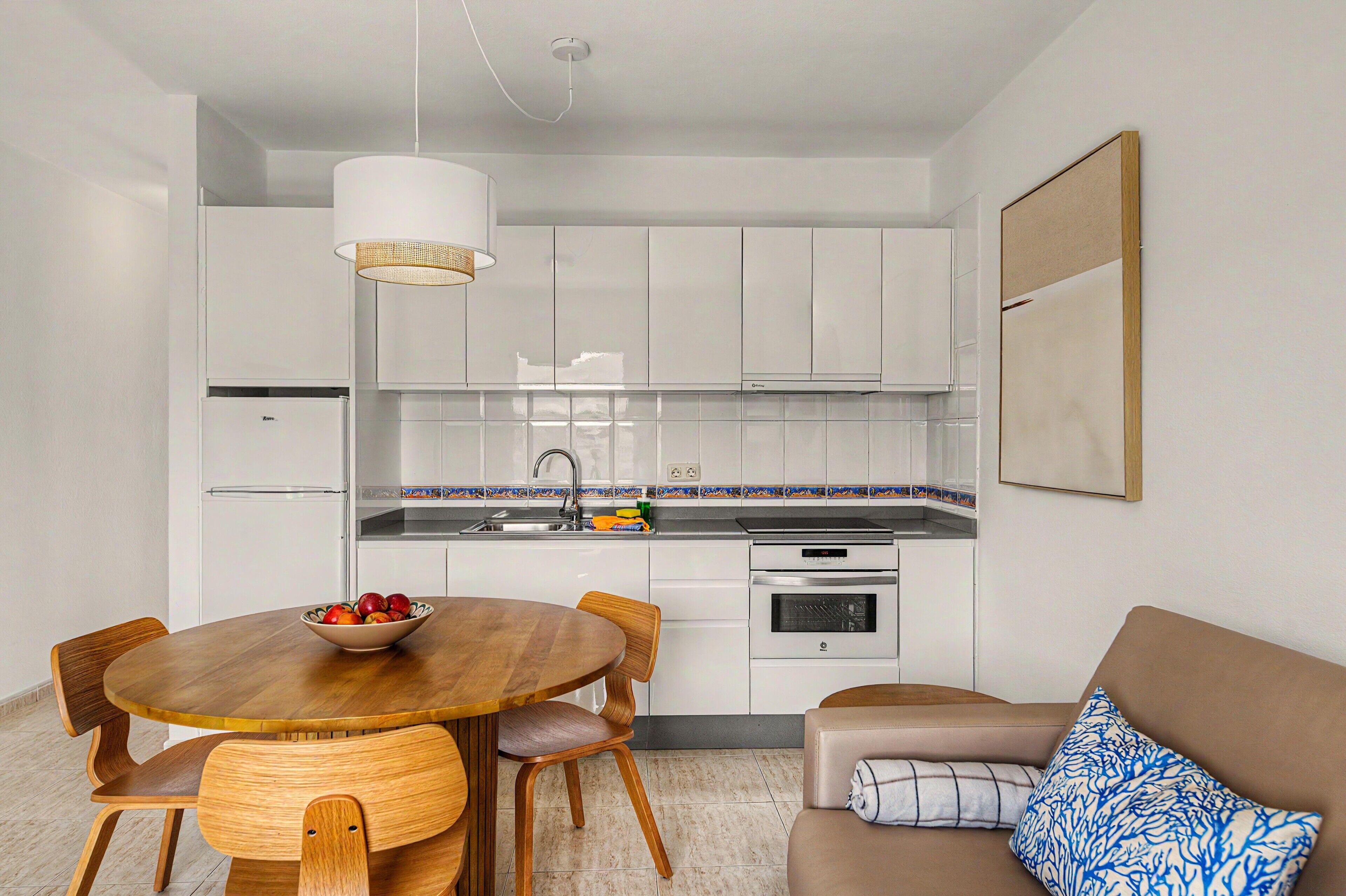 Une cuisine moderne avec des meubles blancs, une table à manger avec des chaises et un canapé agrémenté d’un coussin à motifs.