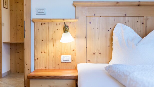 Ein hölzernes Bett mit weißer Bettwäsche, ein Nachttisch mit Schublade, eine Wandleuchte und ein Lichtschalter.