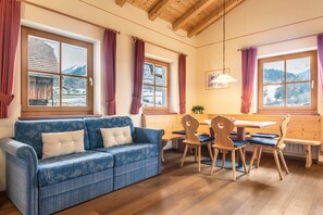 A cozy room with a blue patterned sofa, wooden dining table, and chairs, large windows with curtains, and a wooden ceiling. - Farm Vacations - "Fewo Molignon Paalhof" with Mountain View, Wi-Fi & Balcony (Kastelruth)
