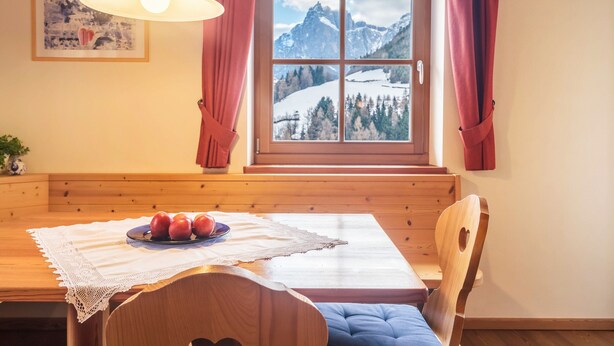Taula de dinar de fusta amb una estovall de punt de gots, un plat d'apples i una vista de muntanyes nevades a través d'una finestra amb cortines vermelles.