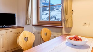 Une cuisine avec des meubles en bois, un téléviseur, une fenêtre donnant sur un paysage enneigé et un saladier de fruits sur la table.