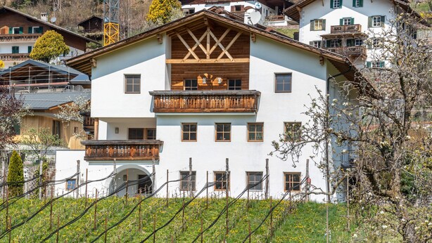 Un immeuble à plusieurs étages doté d’un balcon, un vignoble au premier plan et d’autres bâtiments en arrière-plan.