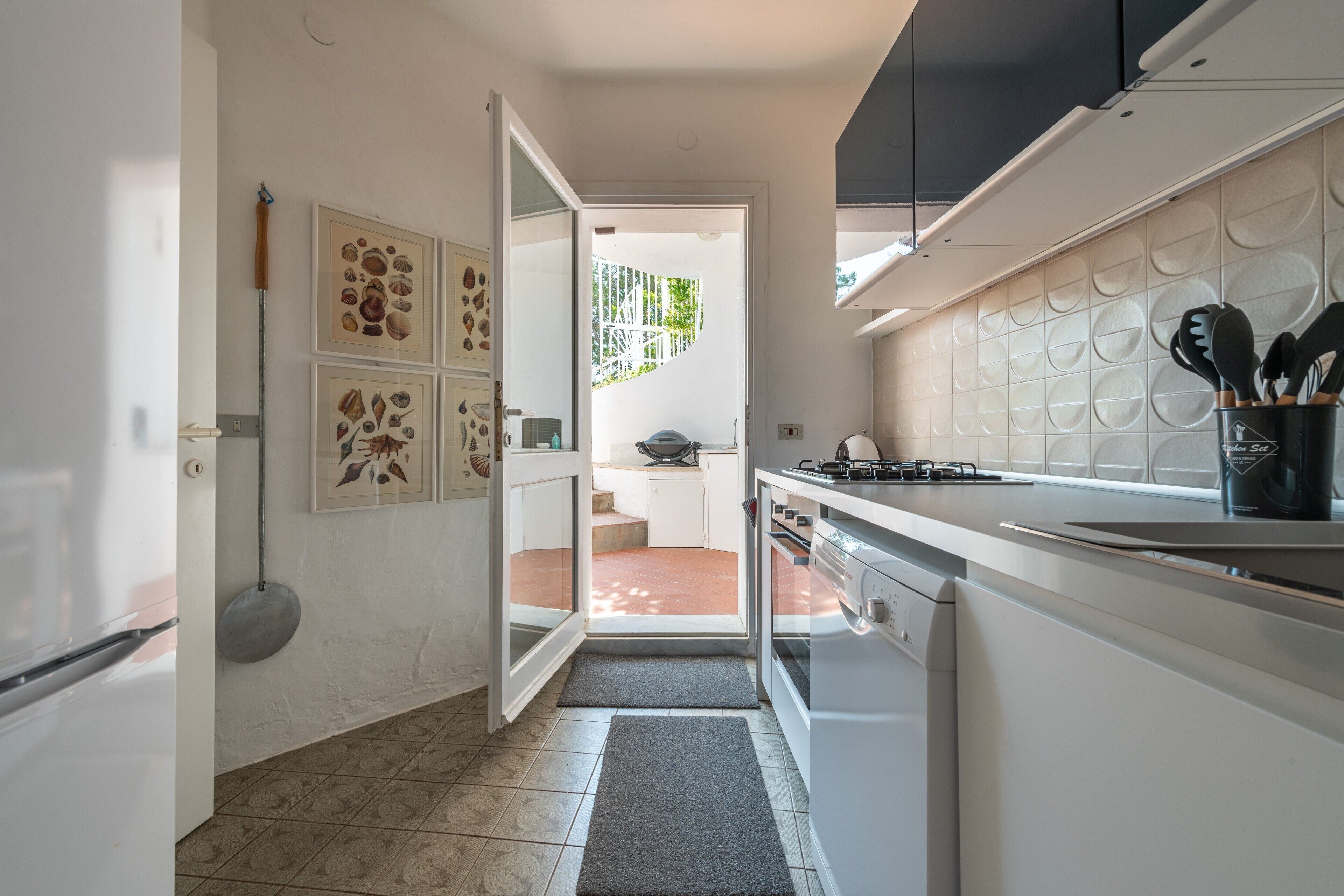 Cucina moderna con mobili bianchi, piano cottura e lavello. Una porta dà accesso a un balcone con vista su aree verdi.