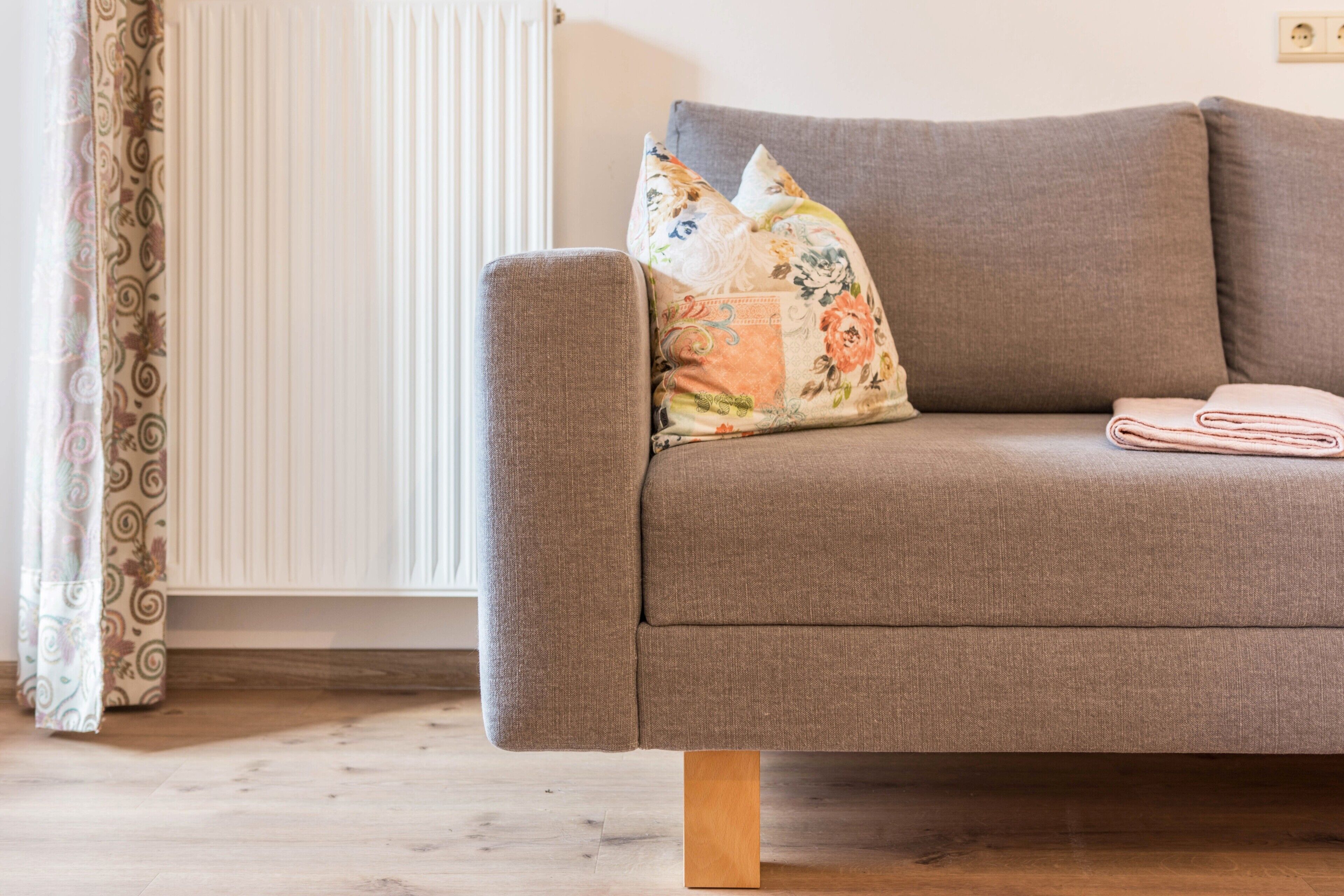 Un sofá gris con cojines estampados y una manta doblada, patas de madera, un radiador y cortinas con diseño.