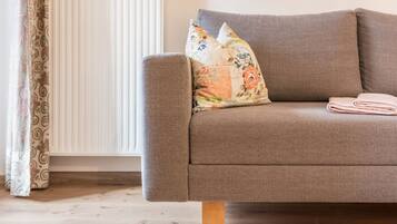 Un sofá gris con cojines estampados y una manta doblada, patas de madera, un radiador y cortinas con diseño.
