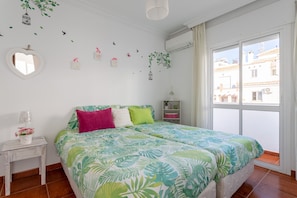 Un dormitorio con cama, mesita de noche, espejo y ventana con vistas a edificios.