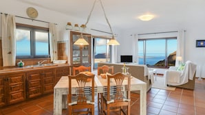 Una cucina con mobili in legno, un tavolo da pranzo con sedie, un lampadario e ampie finestre con vista mare.