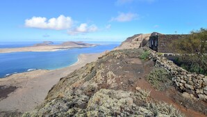 Paesaggio costiero con un muro in pietra, terreno roccioso e vista sul mare e colline in lontananza.