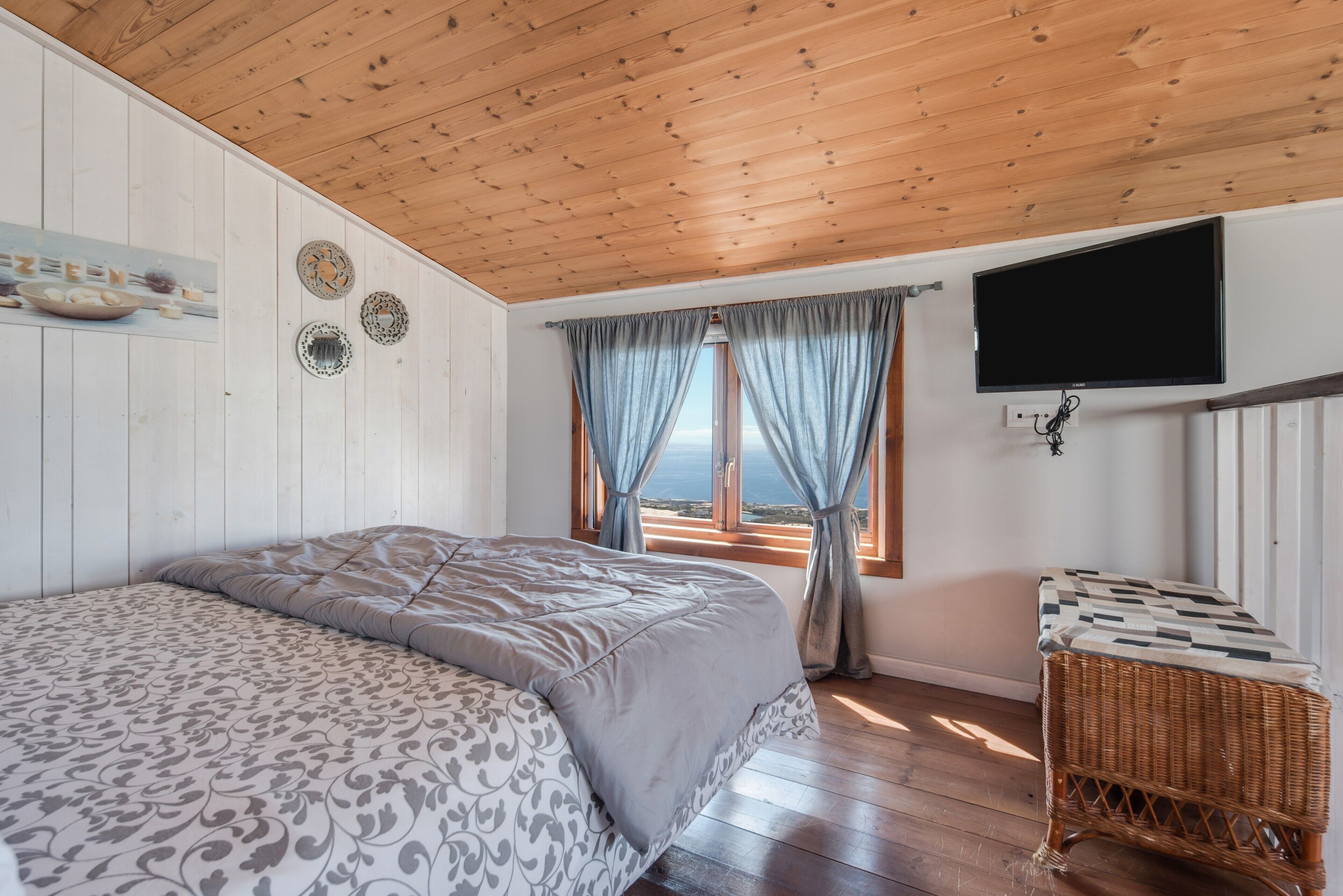 Een slaapkamer met een houten plafond, een bed met beddengoed met een patroon, een aan de muur bevestigde televisie en uitzicht door een raam met gordijnen.
