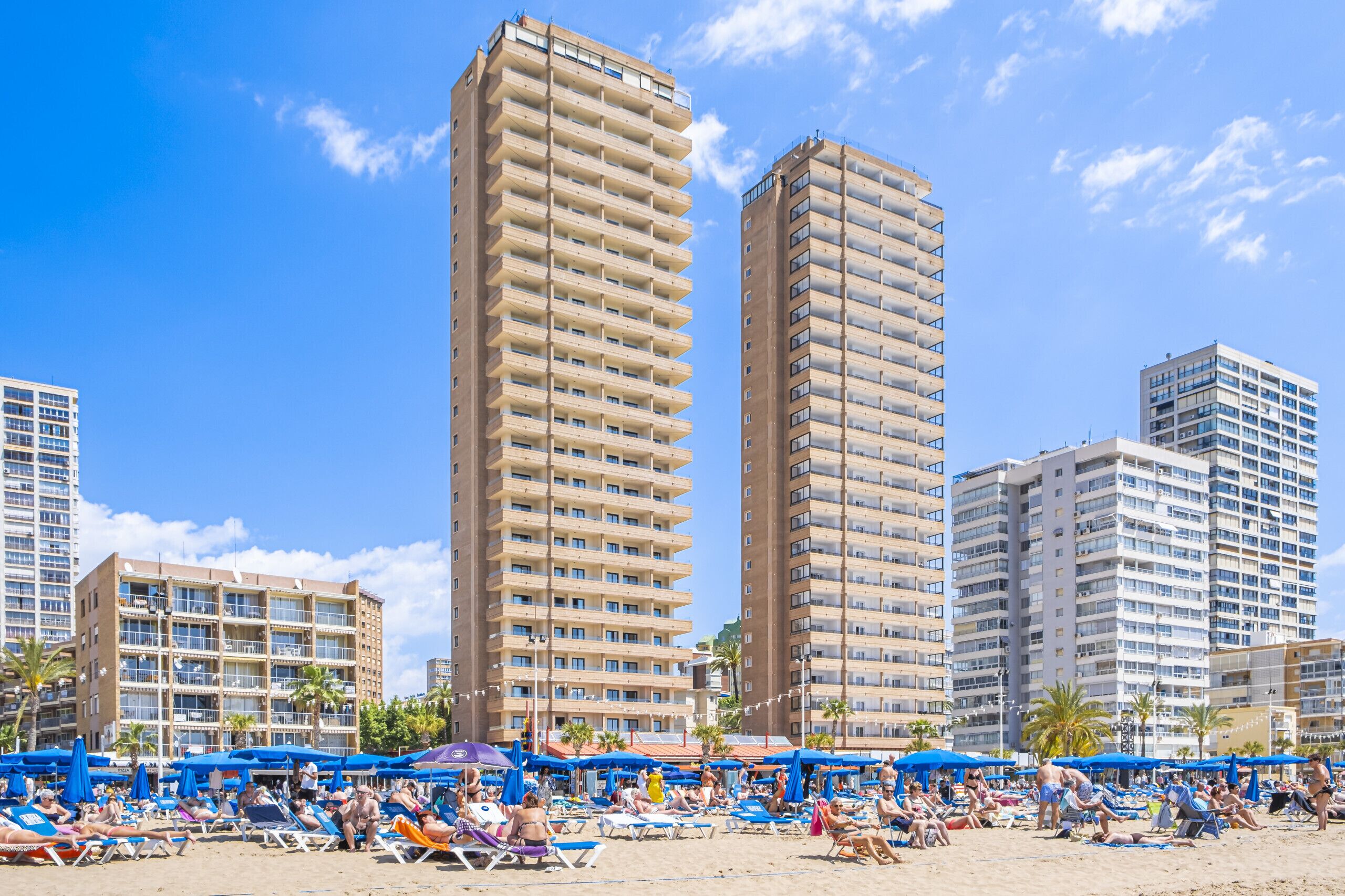 A beach with sun loungers and umbrellas, surrounded by tall residential buildings.