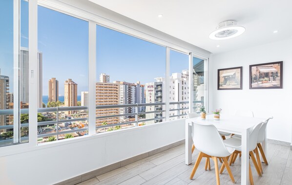 Habitación de hotel moderna con ventanas amplias que ofrecen vistas a la ciudad, una mesa de comedor blanca con sillas y dos cuadros en la pared.