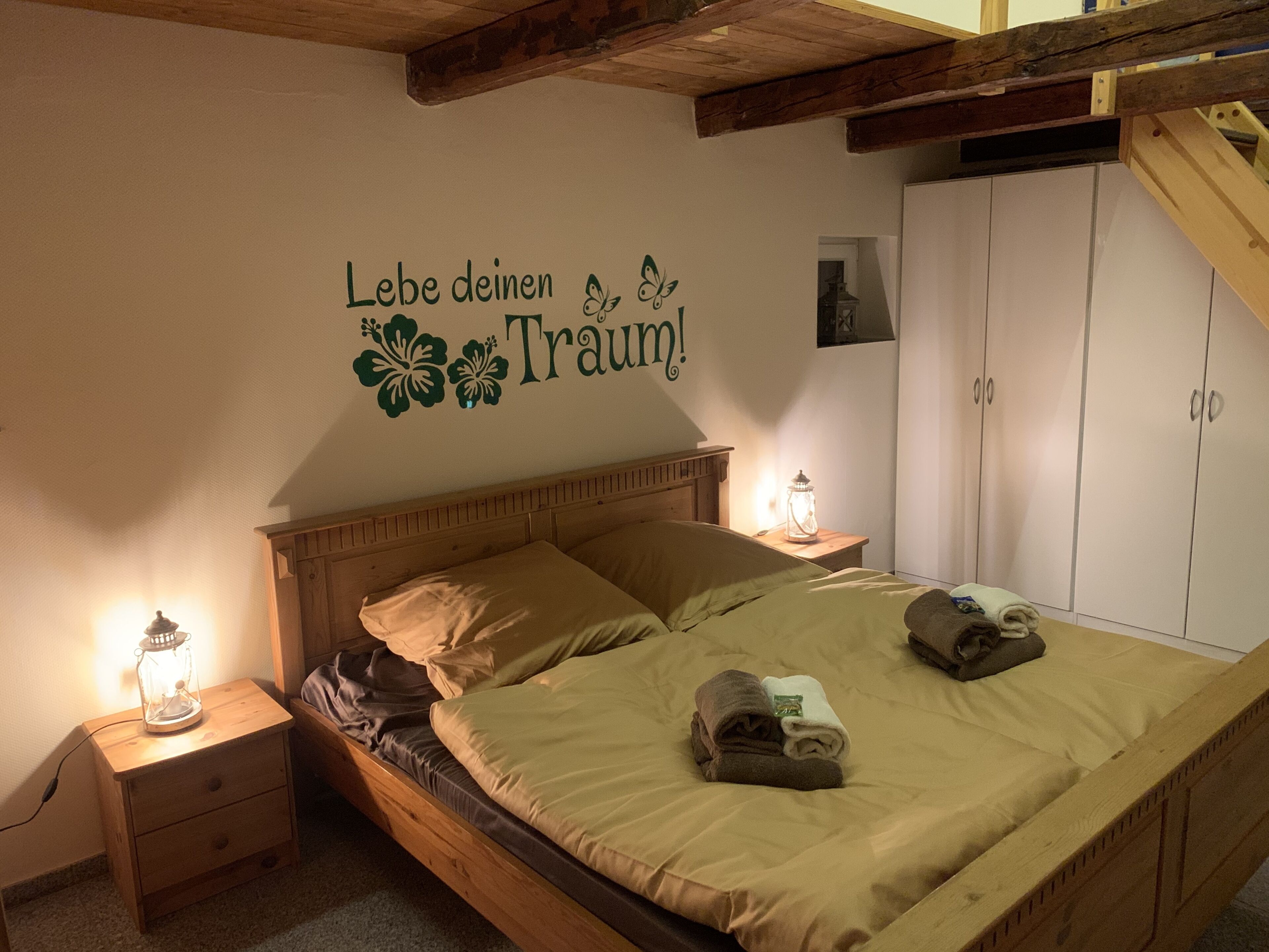 A bedroom with a wooden bed, two nightstands, a wall with a decorative phrase, a wardrobe, and a hanging light fixture.