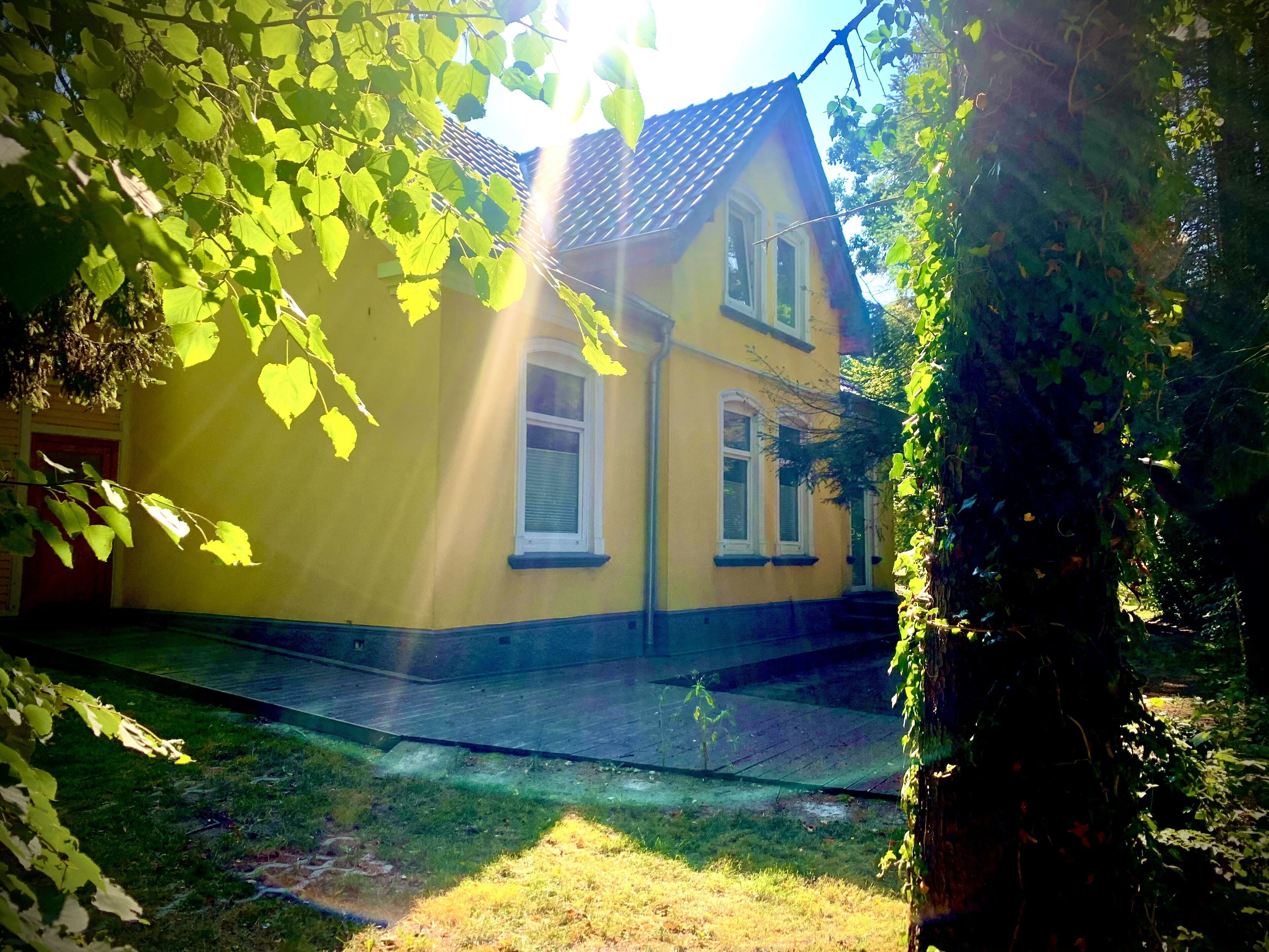 A yellow house with a tiled roof, surrounded by greenery and a paved area.