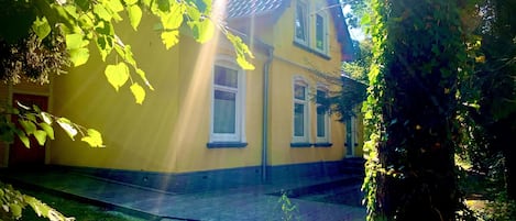 A yellow house with a tiled roof, surrounded by greenery and a paved area.