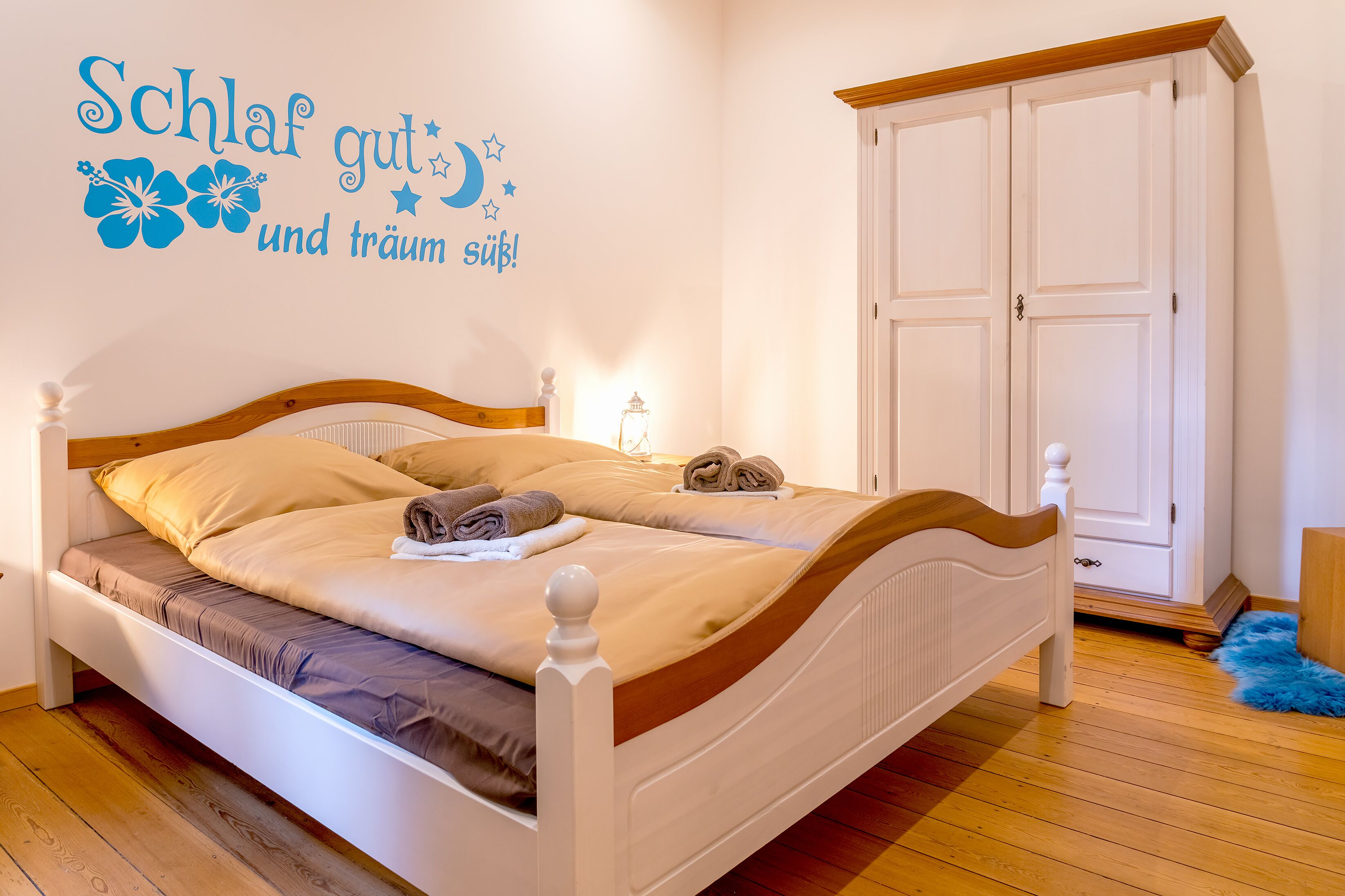 A neatly made bed with a wooden headboard, a wall decoration with text and flowers, and a wardrobe in the background.