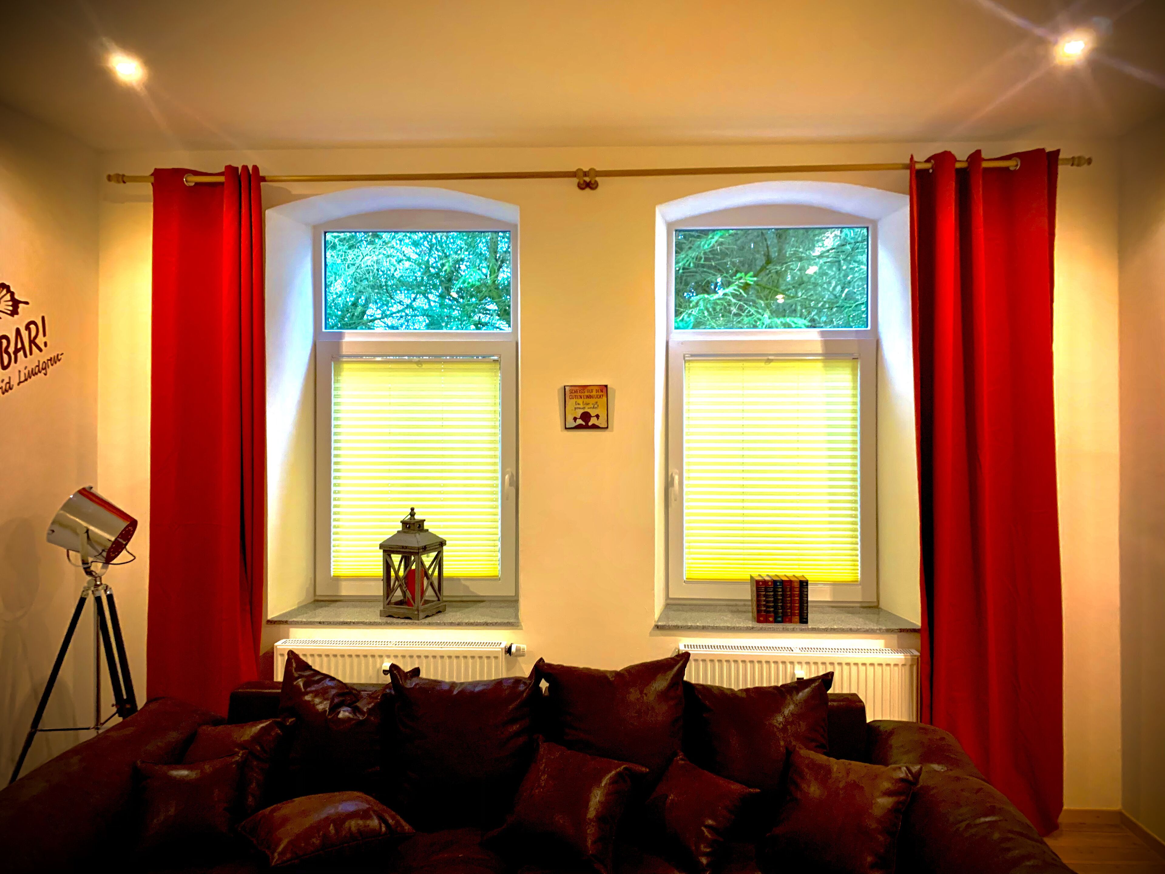 Ein gemütliches Wohnzimmer mit einer Ledersofa, zwei Fenstern mit roten Vorhängen, einem kleinen Tisch mit Büchern und einer Dreibeinlampe.