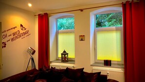 A cozy living room with a red sofa, a floor lamp, and a wall with a quote. There are windows with blinds and a view of greenery outside. - Seasides Villa Kunterbunt (Scharbeutz)