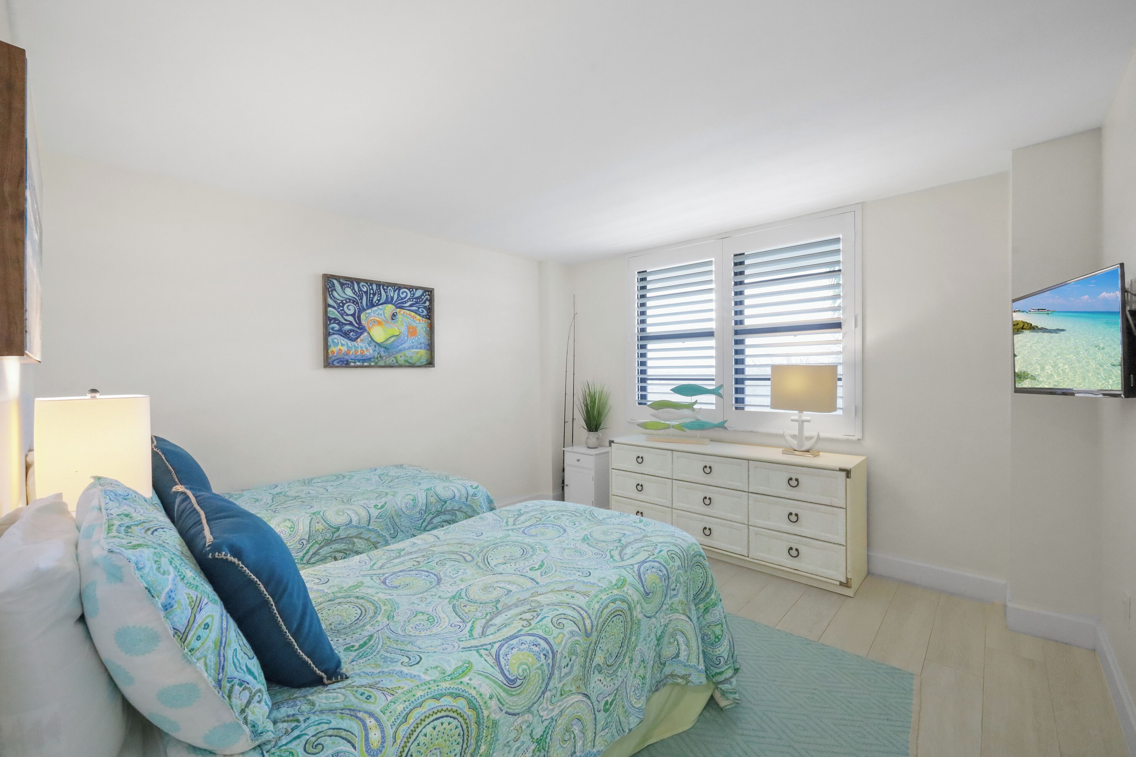 Een slaapkamer met twee bedden, een dressoir, een televisie en een raam met jaloezieën.