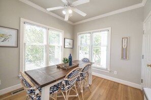 A dining area with a wooden table and four chairs, large windows with blinds, a ceiling fan, and a 'WELCOME' sign on the wall.