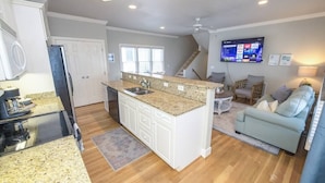 A modern kitchen with granite countertops, white cabinetry, and a central island with a sink. There is a dining area with a table and chairs, and a light blue sofa in the living room.