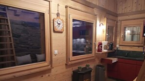 Interior acogedor de una cabaña de madera con pared de piedra, escalera de madera, chimenea y zona de cocina con pileta y armarios rojos.
