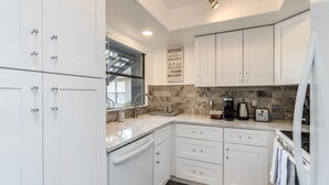 Una cocina moderna con armarios blancos, encimera de mármol y salpicadero de piedra. Se observa una cafetera, un tostador y un lavamanos con grifo.