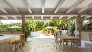 An outdoor patio with a wooden table and chairs, a stone bench, and a pool area in the background. - Luxurious Ganesh Villa (Nicoya)