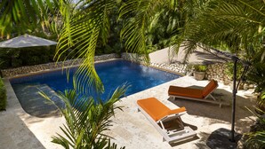 A rectangular swimming pool with a stone border, surrounded by palm trees and a stone wall. There are two lounge chairs with orange cushions and a white umbrella. - Luxurious Ganesh Villa (Nicoya)