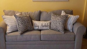 Un sofà gris amb coixins estampats i un coixí amb el text "Stay Awhile", davant d'una paret groga amb un quadre de paisatge emmarcat a la part superior.