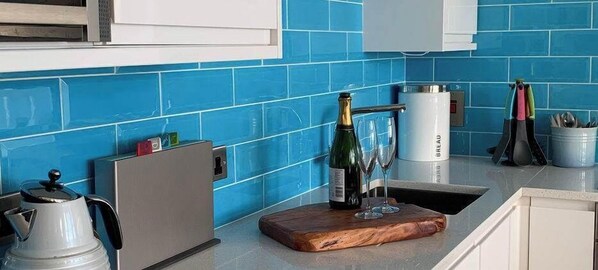 A kitchen with blue tiles, a wooden cutting board, a bottle of champagne, and a coffee maker.
