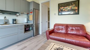 Une cuisine moderne avec des meubles gris, un réfrigérateur en acier inoxydable et une machine à café. On y trouve également un canapé en cuir rouge et une œuvre d’art encadrée accrochée au mur.