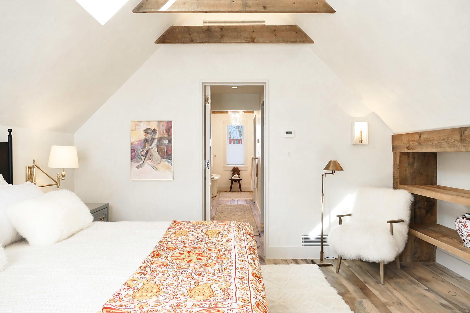 A bedroom with a sloped ceiling, a bed with a patterned bedspread, a white furry chair, a wooden shelf, and a painting on the wall.